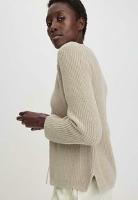 Suéter de punto en beige apagado, con una superficie texturizada, mangas largas y aberturas en los costados del dobladillo para facilitar el movimiento.