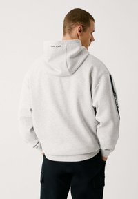 Sudadera con capucha gris que presenta un logo en la espalda y una franja oscura en las mangas, hecha de un tejido suave y texturizado.