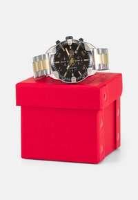 Montre au cadran noir avec un bracelet en acier inoxydable argenté et doré, présentée sur une boîte rouge. Elle dispose de sous-cadrans chronographes et de marquages horaires en relief.