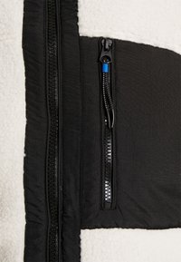 Giacca in pile nera con un pannello testurizzato crema, caratterizzata da un design a doppia zip e una piccola tasca con zip con accenti blu.