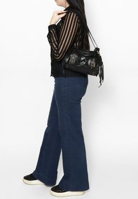 Borsa a tracolla in pelle nera con hardware dorato. Il modello indossa una maglietta a strisce nere e jeans in denim svasati, abbinati a sneakers nere.