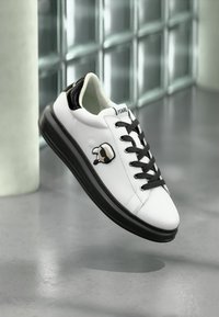 Bijele kožne tenisice s crnim detaljima, s crnom logom oznakom, okruglim vrhom i vezanjem. Gumeni potplat s glatkom teksturom.