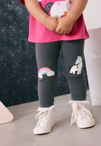 Leggings grises con apliques de arcoíris y unicornios en las rodillas, combinadas con zapatillas blancas de caña alta con detalles de encaje. Camiseta de color rosa brillante.