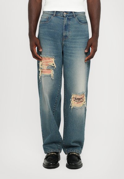 Lichtblauwe denim jeans met een relaxte pasvorm, voorzien van grote, versleten patches op de knieën en rafelige randen. Draagt met zwarte instapschoenen.