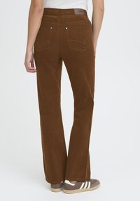Brown corduroy trousers with a straight leg, featuring back pockets, visible stitching, and a branded waistband. Worn with grey sneakers.