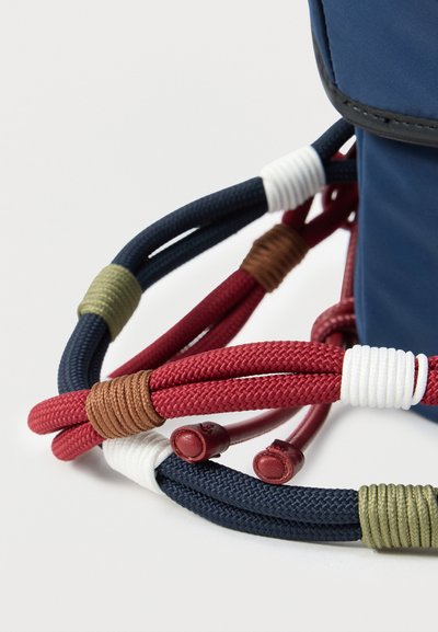 Εγγύς λήψη πλεγμένων κορδονιών κόκκινου και ναυτικού μπλε χρώματος με περιτυλίγματα από χρωματιστές κλωστές, στερεωμένα σε ένα μπλε υφασμάτινο αντικείμενο.
