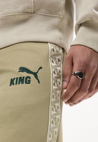 Μπεζ παντελόνι φόρμας Puma King με λογότυπο και σχεδιασμένη πλευρική ρίγα, φορεμένο με μπεζ φούτερ· κοντινή εικόνα χεριού που φορά ασημένιο δαχτυλίδι.