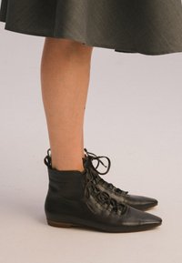 Stivali neri in pelle fino alla caviglia con punta affusolata, dotati di lacci sulla parte anteriore e una texture liscia, posati su un tacco basso marrone.