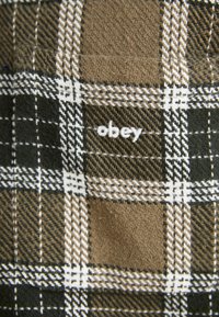 Hnedo-zelený kockovaný materiál s bielymi pruhmi, ktorý obsahuje vyšívanú nálepku "obey". Mäkká textúra, tlmená farebná paleta a tradičný kockovaný vzor.