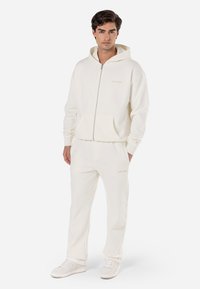 Sweatshirt à capuche blanche zippée avec poches kangourou et poignets côtelés, associé à un pantalon de jogging assorti avec un détail de logo. Texture lisse.