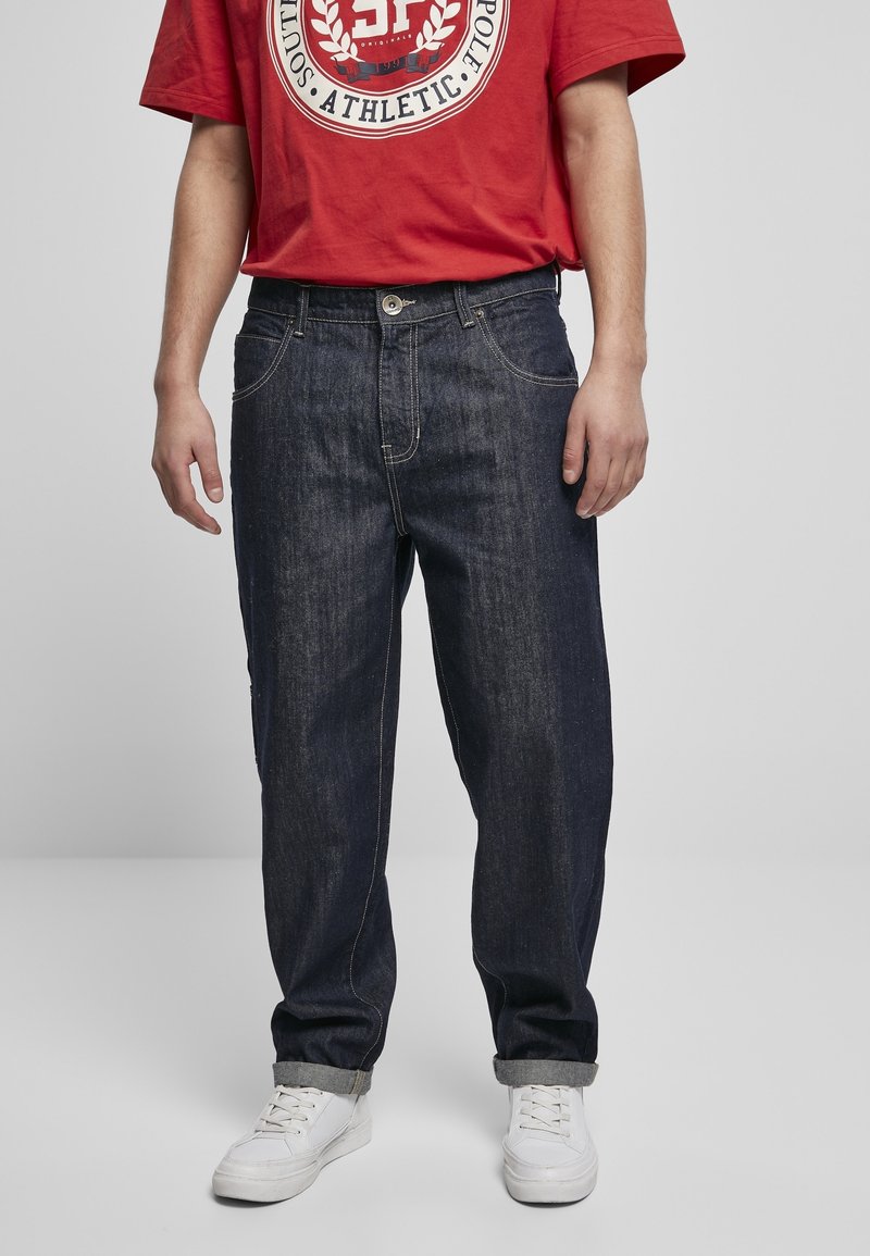 Homme portant un jean bleu foncé retroussé, des baskets blanches, et un t-shirt rouge avec un texte graphique blanc et marine sur un fond gris uni.