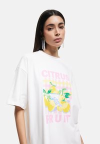 T-shirt blanc surdimensionné avec un graphique coloré de citron et des feuilles stylisées sur un fond à motif de carreaux pastel. Manches courtes et décontractées.