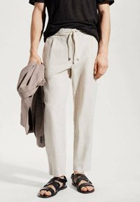 Pantalon en lin beige clair avec une ceinture à cordon, associé à un blazer gris posé sur le bras et des sandales noires à brides.