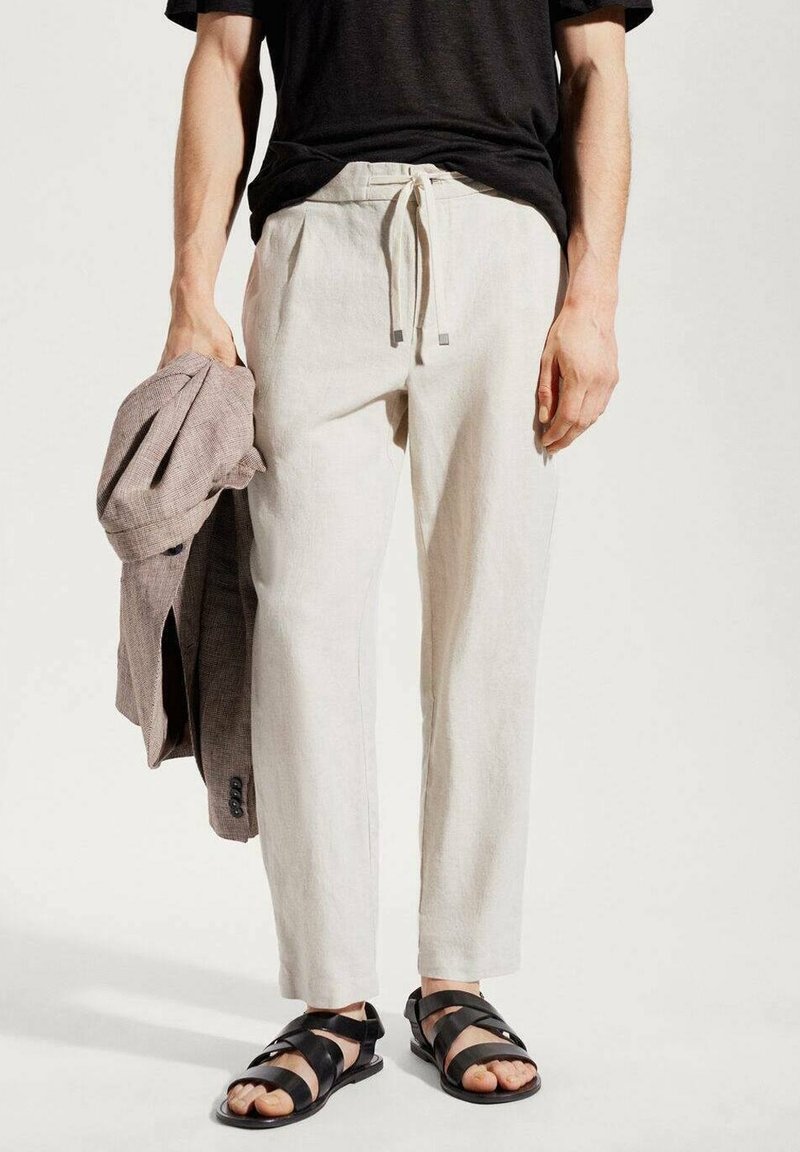 Pantalon en lin beige clair avec une ceinture à cordon, associé à un blazer gris posé sur le bras et des sandales noires à brides.
