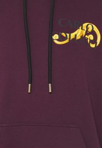 Sudadera con capucha color burdeos con cordones negros y texto "CARLO" con detalles en dorado y un remolino decorativo. Tela suave y bolsillo tipo canguro.