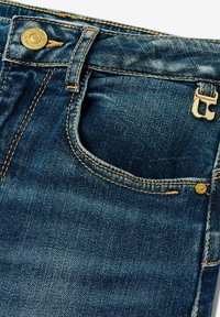 Gros plan sur une poche avant de jeans en denim bleu foncé avec un bouton en laiton visible, une rivet et un surfil orange contrastant.