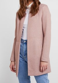 Ljusrosa skräddarsydd kappa med långa ärmar, öppen fram, och två sidofickor, bärs över en vit topp och blå denimjeans. Texturerat tyg.