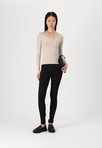 Haut beige à manches longues côtelé avec détails en boutons, associé à un legging noir ajusté et des chaussures noires. Modèle tenant un sac noir.