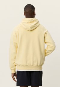 Κίτρινο φούτερ με κουκούλα και κορδόνι, μανίκια ραγκλάν και ριμπ μανσέτες. Μαλακό, υφασμένο ύφασμα με υφή; άνετη εφαρμογή. Φορεμένο με μαύρο σορτς.