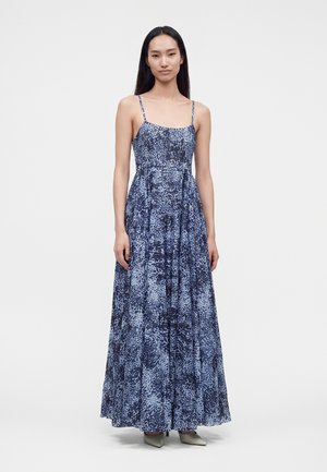 Frau mit langem dunklem Haar, die ein ärmelloses blau-schwarzes gemustertes Maxikleid und hellfarbene spitze Schuhe trägt, steht vor einem weißen Hintergrund.