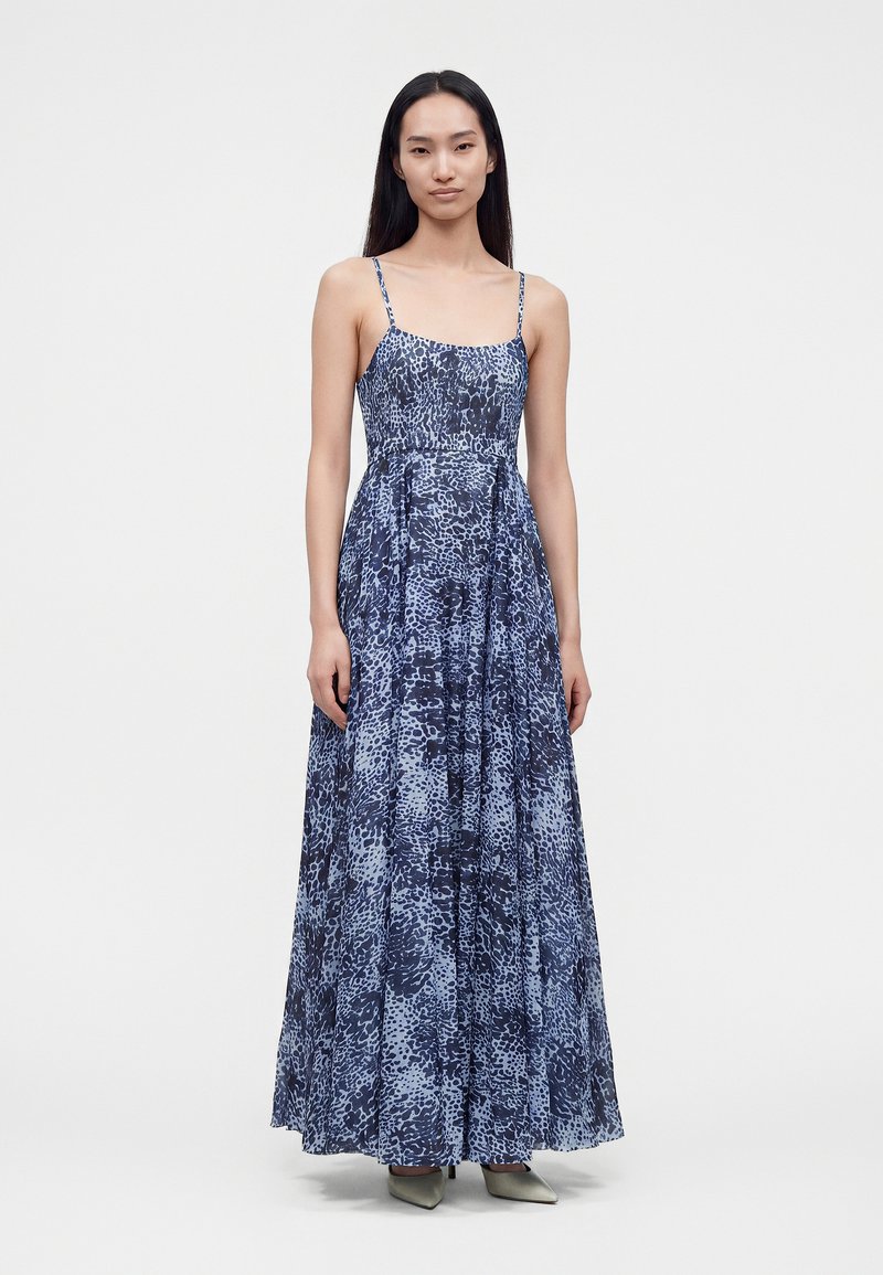 Femme aux longs cheveux foncés portant une robe longue sans manches à motifs bleus et noirs et des chaussures pointues de couleur claire, debout devant un fond blanc.