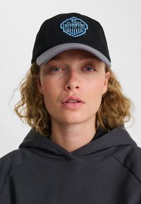 Svart basebollkeps med blå broderad logotyp som har en skölddesign. Kepsen har en grå undersida på skärmen och en avslappnad passform.