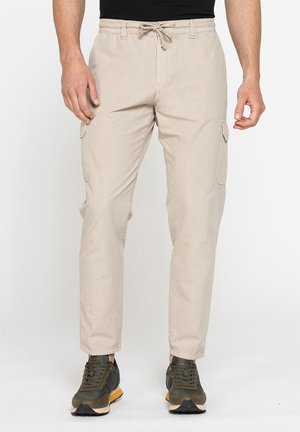 Uomo con pantaloni cargo beige con tasche laterali e sneaker verdi, in piedi davanti a uno sfondo chiaro e uniforme.