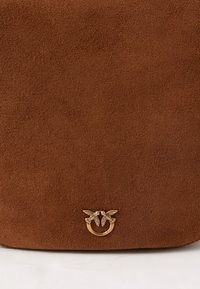 Καφέ σουέτ τσάντα χειρός με λεία υφή και καμπύλη μορφή. Διαθέτει χρυσό έμβλημα με δύο πουλιά και κυκλική σχεδίαση στο κάτω μέρος.