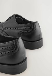 Czarne skórzane eleganckie buty wyróżniające się gładką teksturą, zdobieniem w stylu brogue i okrągłym noskiem. Obcas ma niski profil.