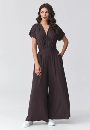 Kobieta stoi z ręką w kieszeni, ubrana w ciemnobrązowy kombinezon z szerokimi nogawkami i dekoltem w serek oraz białe trampki na szarym tle.