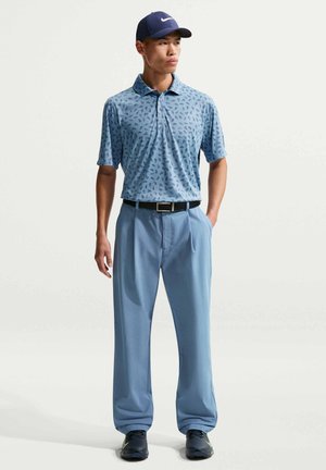 Homme portant un polo à motifs bleu, un pantalon bleu clair, une ceinture noire, une casquette marine et des baskets noires, debout avec une main dans la poche.