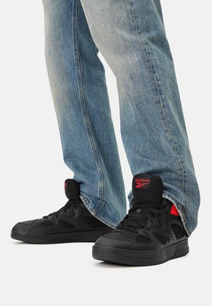 Czarne sneakersy Reebok z czerwonymi akcentami, o teksturowanej cholewce i średniej wysokości kroju, zestawione z jasnoniebieskimi dżinsami.