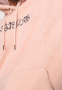 Lichtroze fleece hoodie met een gestructureerd oppervlak. Bevat zwart geborduurde tekst en roze trekkoorden met metalen uiteinden. Voorzak zichtbaar.