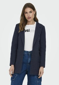 Blazer bleumarin cu model herringbone, cu mâneci lungi, rever cu fante, buzunare laterale și croială dreaptă, purtat peste un tricou alb cu imprimeu și pantaloni din denim.