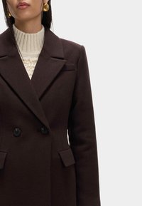 Ciemnobrązowy płaszcz dwurzędowy z ostrymi klapami, o dopasowanym kroju i dwoma przednimi kieszeniami, zestawiony z kremowym swetrem z wysokim kołnierzem.
