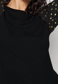 Svart topp med slät textur, med korta ärmar i genomskinligt material prydda med guldprickade mönster.