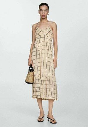 Abito midi beige con motivo a quadri, spalline sottili e orlo a più livelli. La modella tiene una borsa intrecciata con manico nero e indossa sandali neri con lacci.