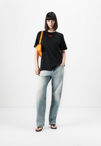 Vrouw die tegen een witte achtergrond staat, gekleed in een zwart T-shirt met een rood logo, lichte vervaagde spijkerbroek, zwarte sandalen en een oranje schoudertas.