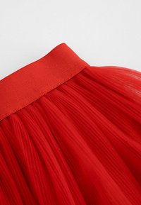Czerwona spódnica tiulowa z szerokim, teksturowanym elastycznym paskiem w talii. Warstwy miękkiego, przezroczystego materiału tworzą objętość i ruch.