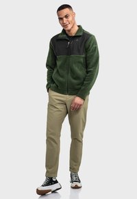 Grüne Fleecejacke mit schwarzem oberen Bereich, Reißverschluss und Seitentaschen; kombiniert mit beigen Hosen und schwarzen Sneakers.