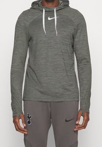 Haut à capuche gris à manches longues en tissu texturé, avec un logo Nike blanc sur la poitrine et des cordons de serrage réglables sur la capuche.