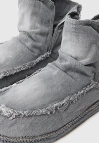 Szare dżinsowe buty typu slip-on o luźnym kroju, z postrzępionymi brzegami i kontrastującymi szwami, charakteryzujące się miękką, teksturowaną powierzchnią.