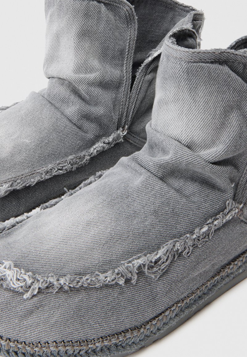 Scarpe slip-on in denim grigio con una vestibilità rilassata, bordi sfilacciati e cuciture a contrasto, caratterizzate da una superficie morbida e testurizzata.