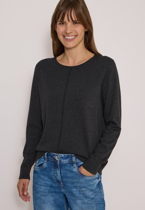 Femme souriante portant un pull gris foncé à manches longues et un jean bleu, avec une main dans la poche sur un fond uni.