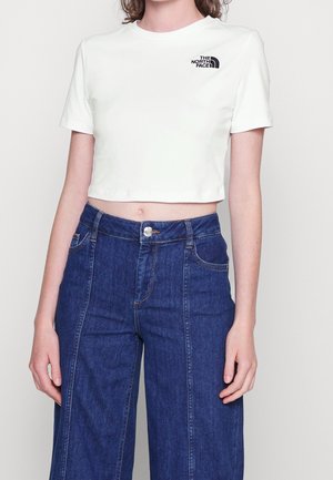 Woman wearing a white cropped t-shirt with The North Face logo and high-waisted wide-leg blue jeans, standing against a plain background.