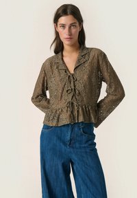Lace blouse in olive green with a peplum hem, featuring a collar, front tie detail, and long sleeves, paired with blue wide-leg jeans.