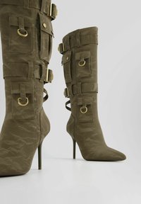 Olivgröna knähöga stövlar med stilettklack, spänne och fickdetaljer, med skrynklig textur och spetsig t å.