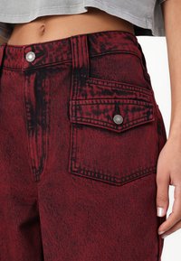 I pantaloni in denim rossi presentano un alto punto vita, una texture strappata, due tasche frontali e dettagli in metallo argentato sul bottone e sulla tasca.