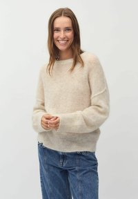Beige, puha, bolyhos anyagból készült kötött pul�óver, laza szabással és ejtett vállakkal. Kék farmernadrággal párosítva, bemutatva egy mindennapi megjelenést.