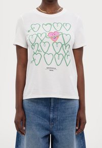 Valkoinen puuvillapaita, jossa on vihreä sydänkuvio ja yksi keskellä oleva vaaleanpunainen sydän ja kasvot. Teksti "VIIMEISET" näkyy graafisen kuvan alla.